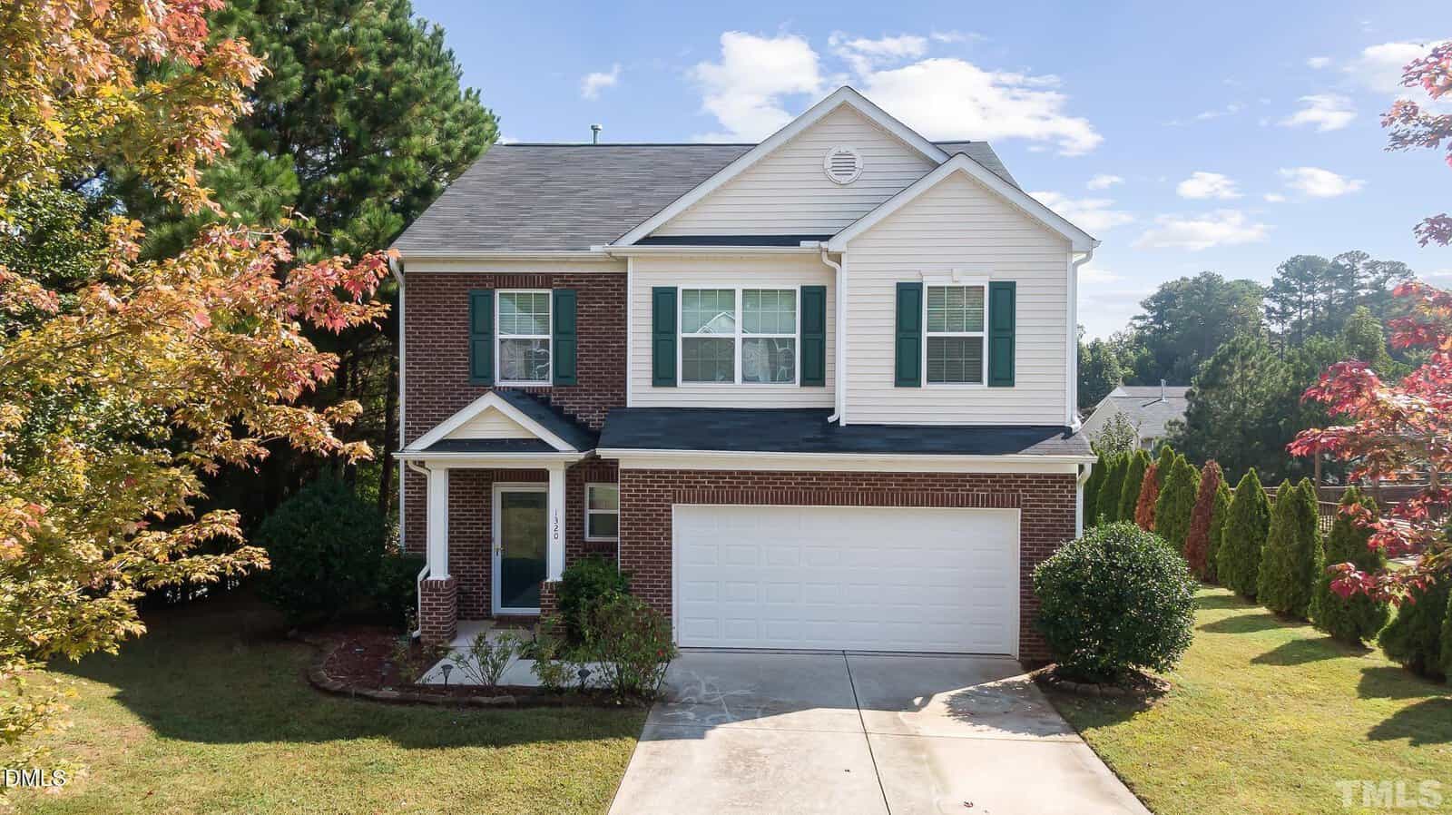A charming two-story suburban house at 1320 Hosmer Court, Wake Forest, with brick and white siding exterior, green shutters, a two-car garage, and a landscaped front yard with trees and shrubs. The sky is mostly clear with a few clouds.
