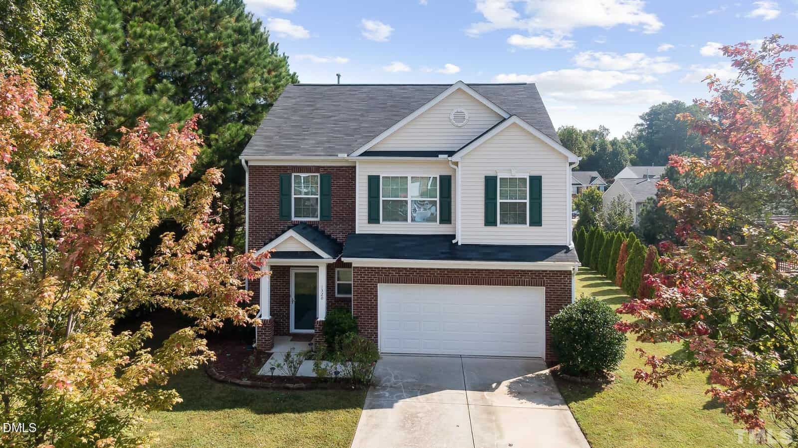 A two-story suburban house with a brick and white siding exterior, double garage, driveway, and green lawn, surrounded by trees with some autumn foliage under a partly cloudy sky.