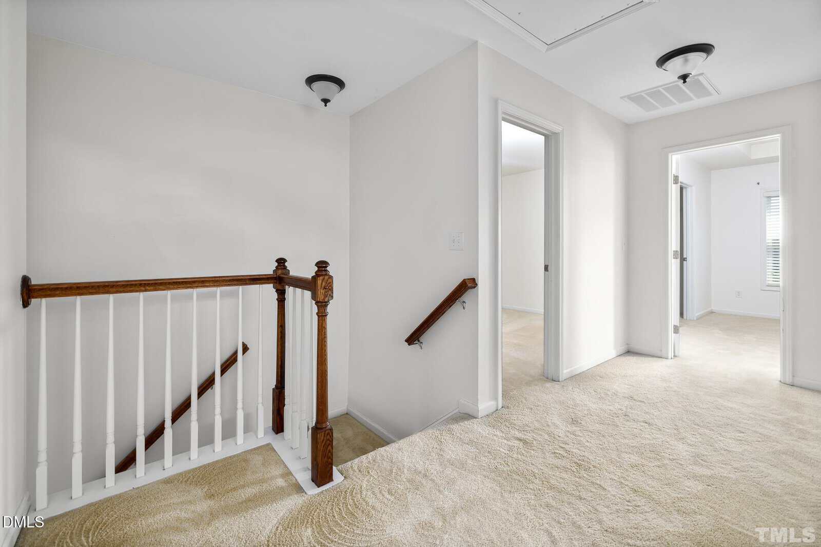 Upstairs hallway with beige carpet, white walls, two ceiling lights, three open doorways, and a wooden handrail leading down the staircase. Natural light enters from windows in adjacent rooms.
