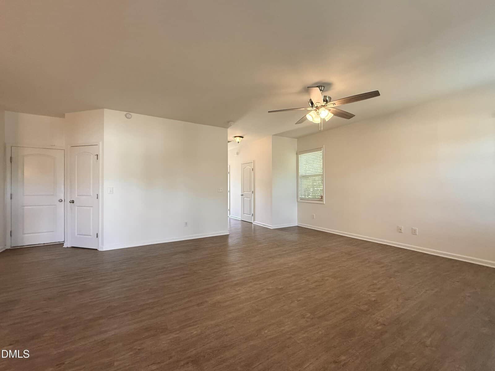 A spacious, empty room with light-colored walls, wood-style flooring, a ceiling fan with lights, and multiple closed white doors. A window with blinds lets in natural light from the right side.