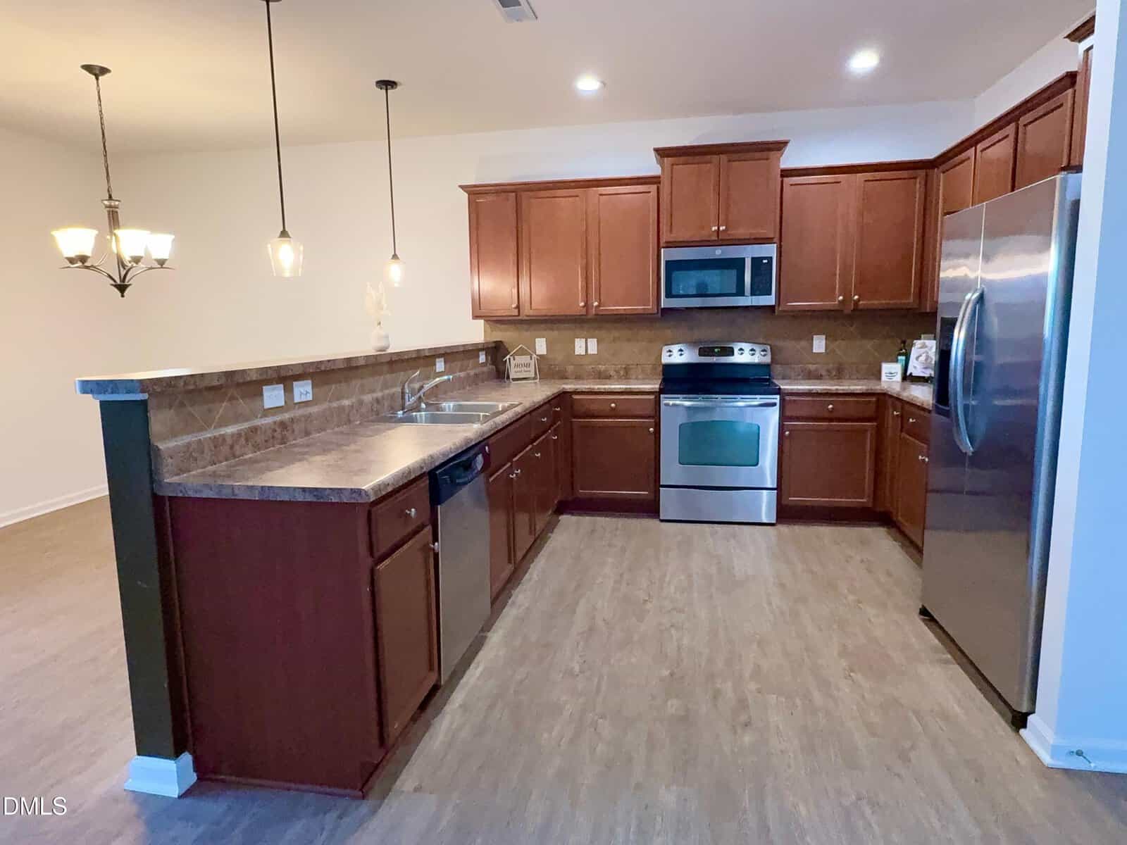A spacious kitchen with wood cabinets, stainless steel appliances including a stove, microwave, and refrigerator, a double sink, dishwasher, brown countertops, and pendant lighting over a breakfast bar.