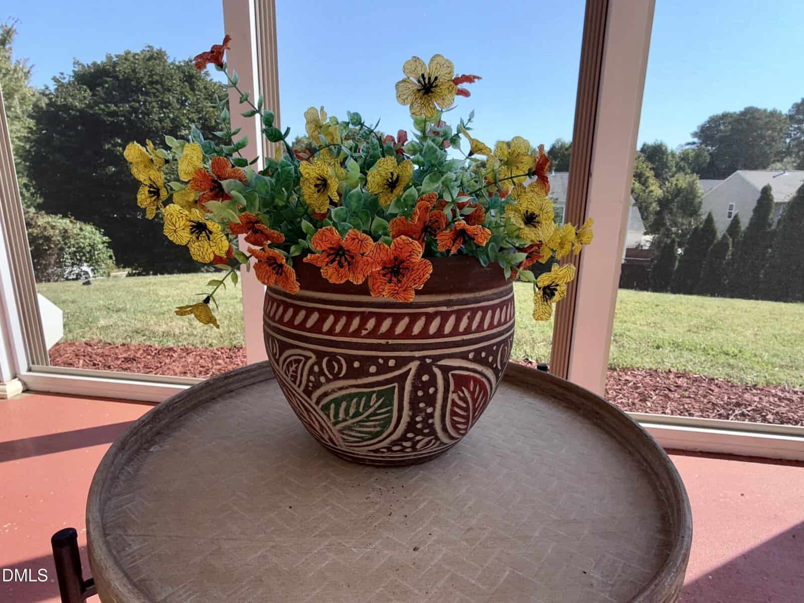 A decorative clay pot with colorful artificial flowers sits on a round table by a window, with a grassy yard and trees visible outside in the sunlight.