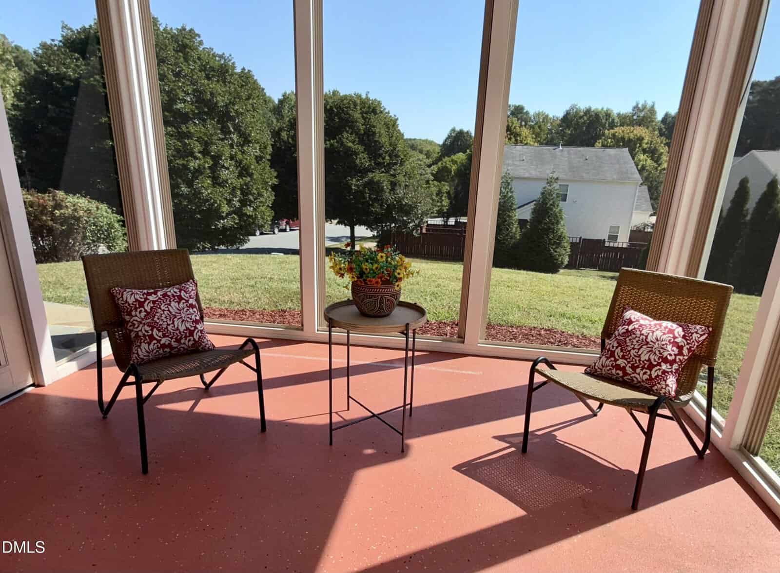 Two wicker chairs with red patterned pillows face each other on a screened-in porch. A small round table with a flower arrangement sits between them. The porch overlooks a grassy yard and nearby houses.