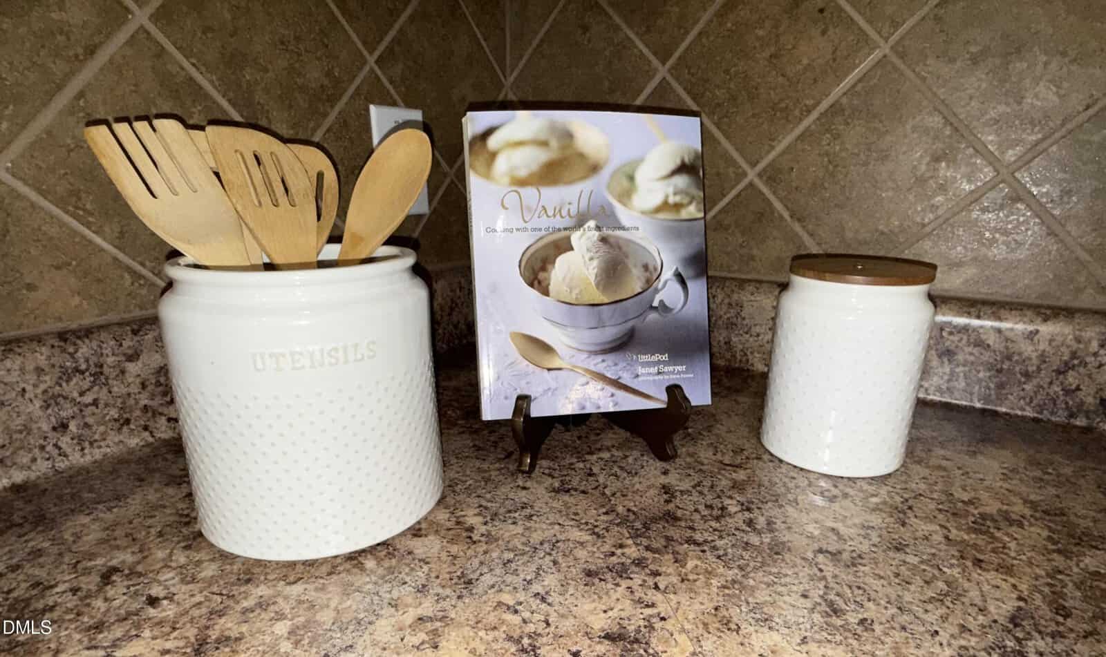 A ceramic utensil holder with wooden cooking tools sits on a kitchen counter next to a matching jar and a vanilla ice cream recipe book displayed on a stand against a tiled backsplash.