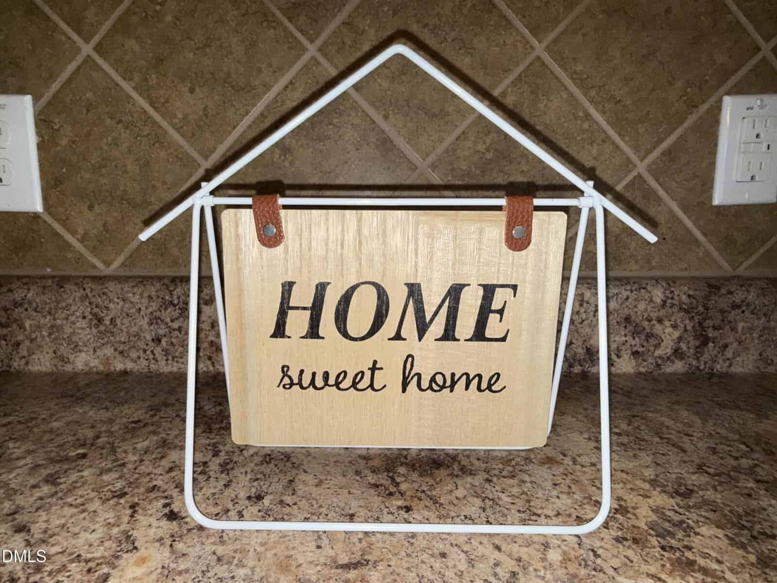 A wooden sign with the words "HOME sweet home" hangs on a white metal frame shaped like a house, set on a brown granite countertop against a tiled backsplash.
