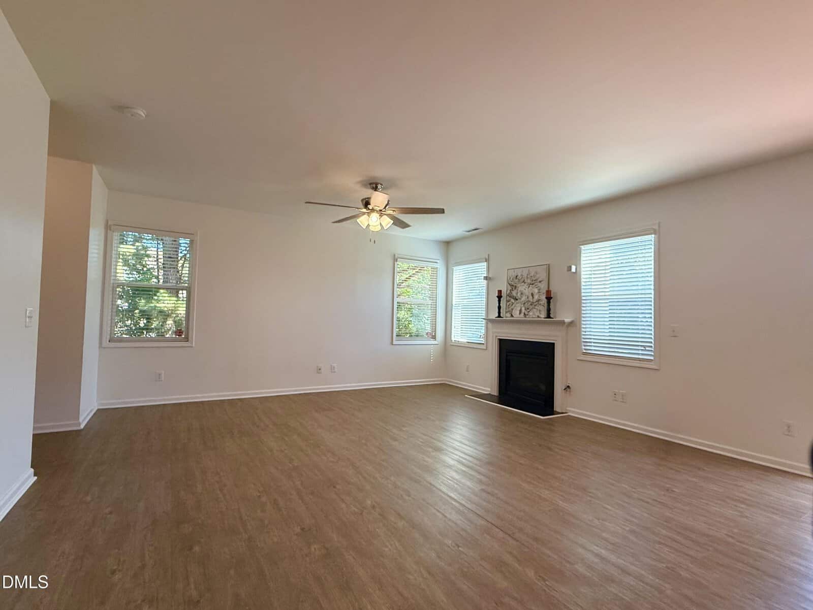 A spacious, empty living room with light-colored walls, wood flooring, three windows, a ceiling fan with lights, and a fireplace with a mantel and decorative artwork. Natural light fills the room.