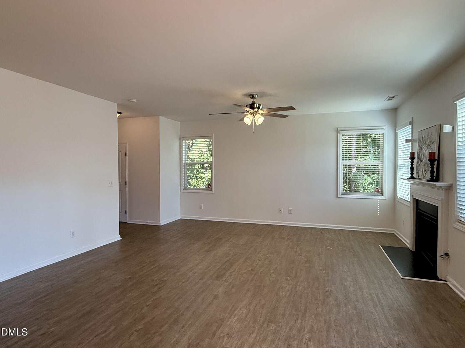 Spacious empty living room with wood flooring, white walls, a ceiling fan with lights, several windows letting in natural light, and a fireplace with a mantle on the right side.
