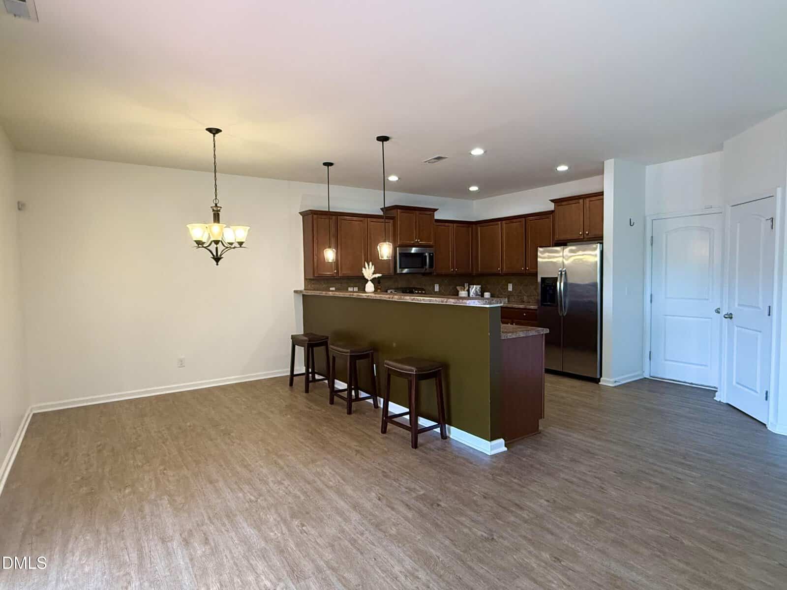 A kitchen with brown cabinets, stainless steel appliances, and a breakfast bar with three stools, next to an empty dining area with wood-style flooring and a chandelier.