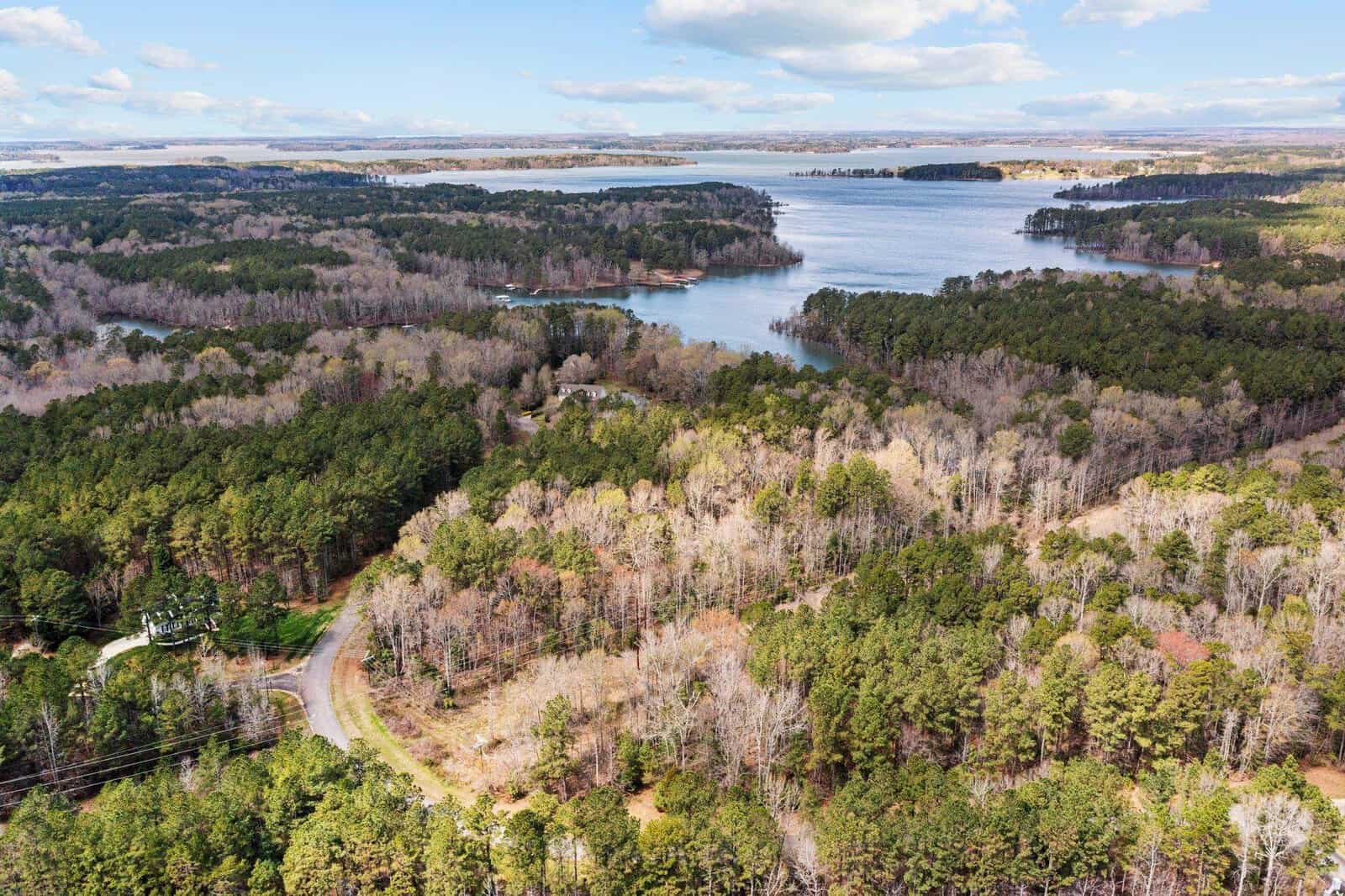 Aerial view of a forested area with a winding road, surrounding a large lake with several inlets and distant tree-covered islands under a partly cloudy sky.
