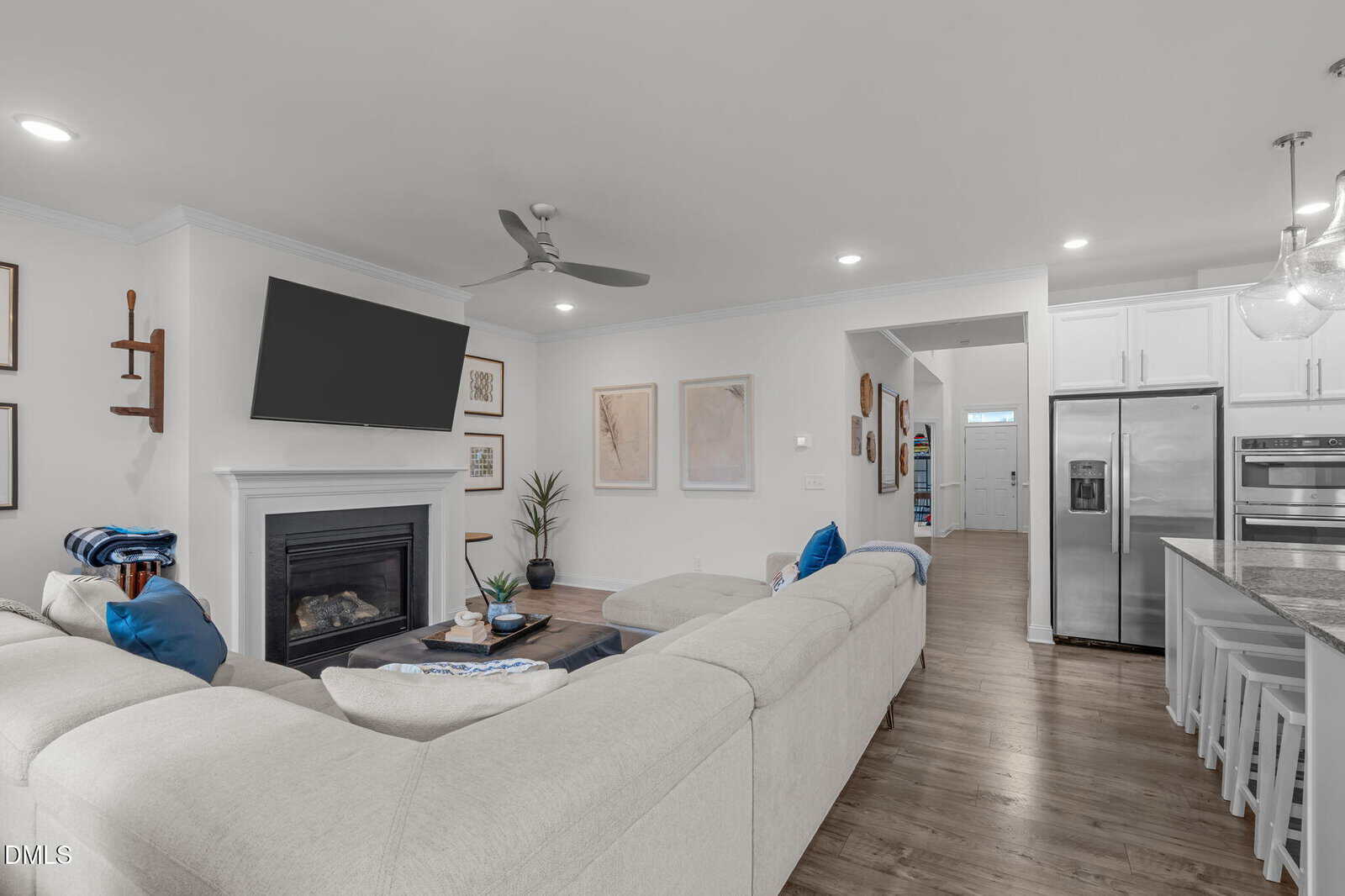 A modern living room with a white sectional sofa, a fireplace below a mounted TV, framed art on the walls, a kitchen area with white cabinets and a stainless steel fridge, and wood flooring throughout.