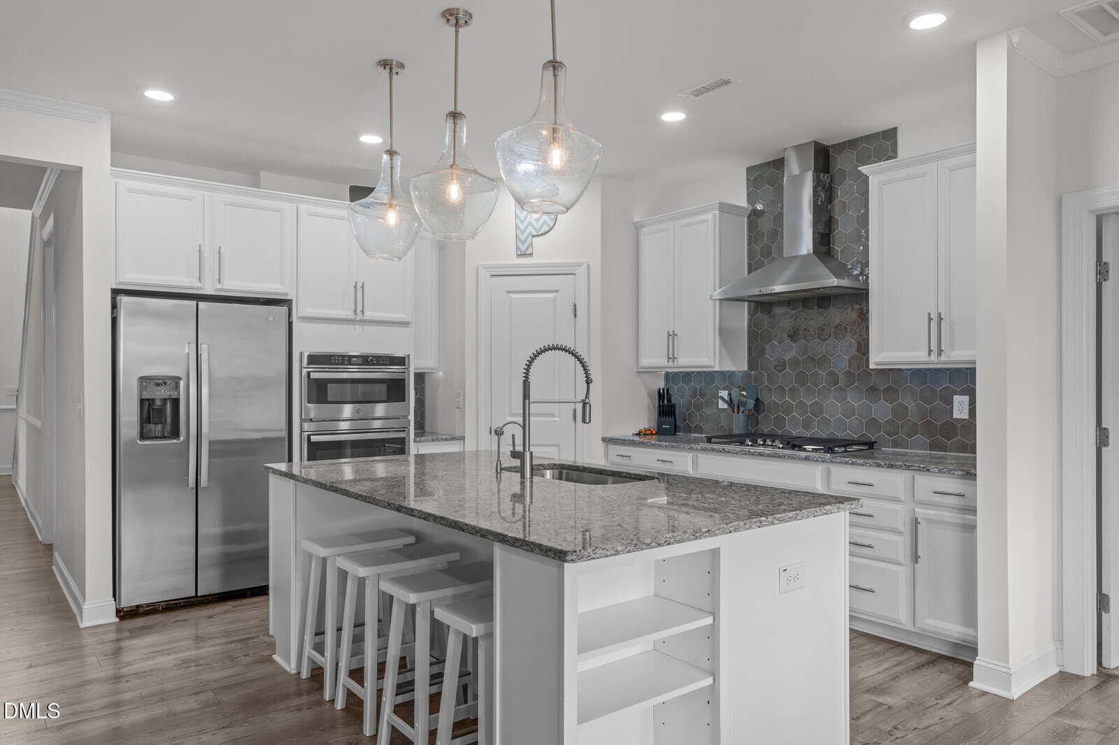 Modern kitchen with white cabinets, stainless steel appliances, a large island with a granite countertop, four white barstools, pendant lighting, and a hexagonal tile backsplash behind the range and vent hood.