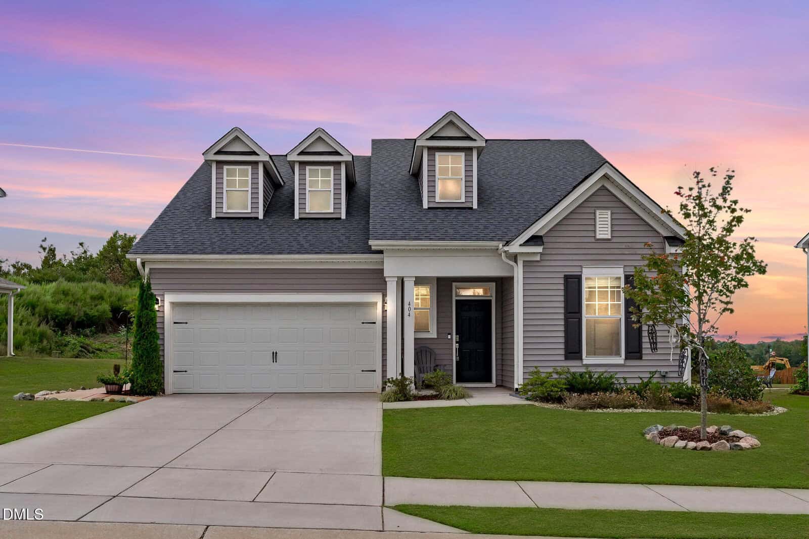 A gray single-family house with white trim, three dormer windows, a two-car garage, a front door with sidelights, landscaped front yard, and a concrete driveway at 404 Porterfield Drive in Youngsville is beautifully photographed at sunset against a pink and purple sky.