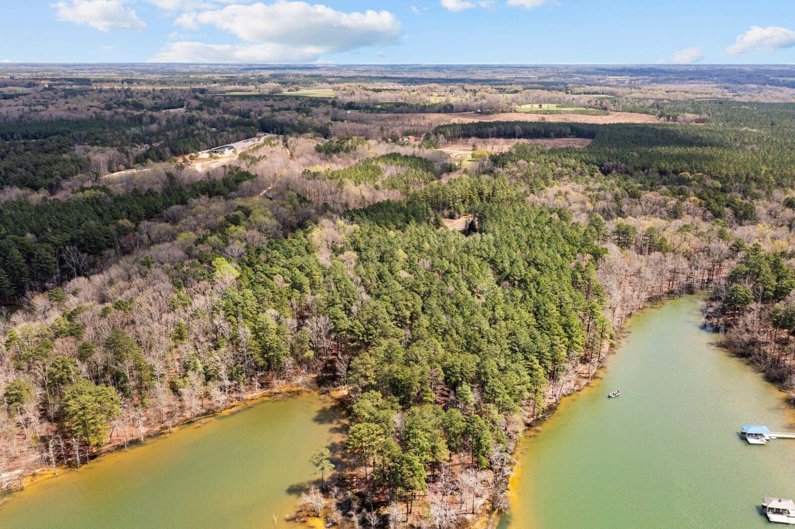 Aerial view of a forested area bordered by two coves of a lake near Manson, with boats on the water, under a partly cloudy sky. Distant fields and more trees are seen in the background, with Lot 19 Key Largo Lane nestled among the greenery.