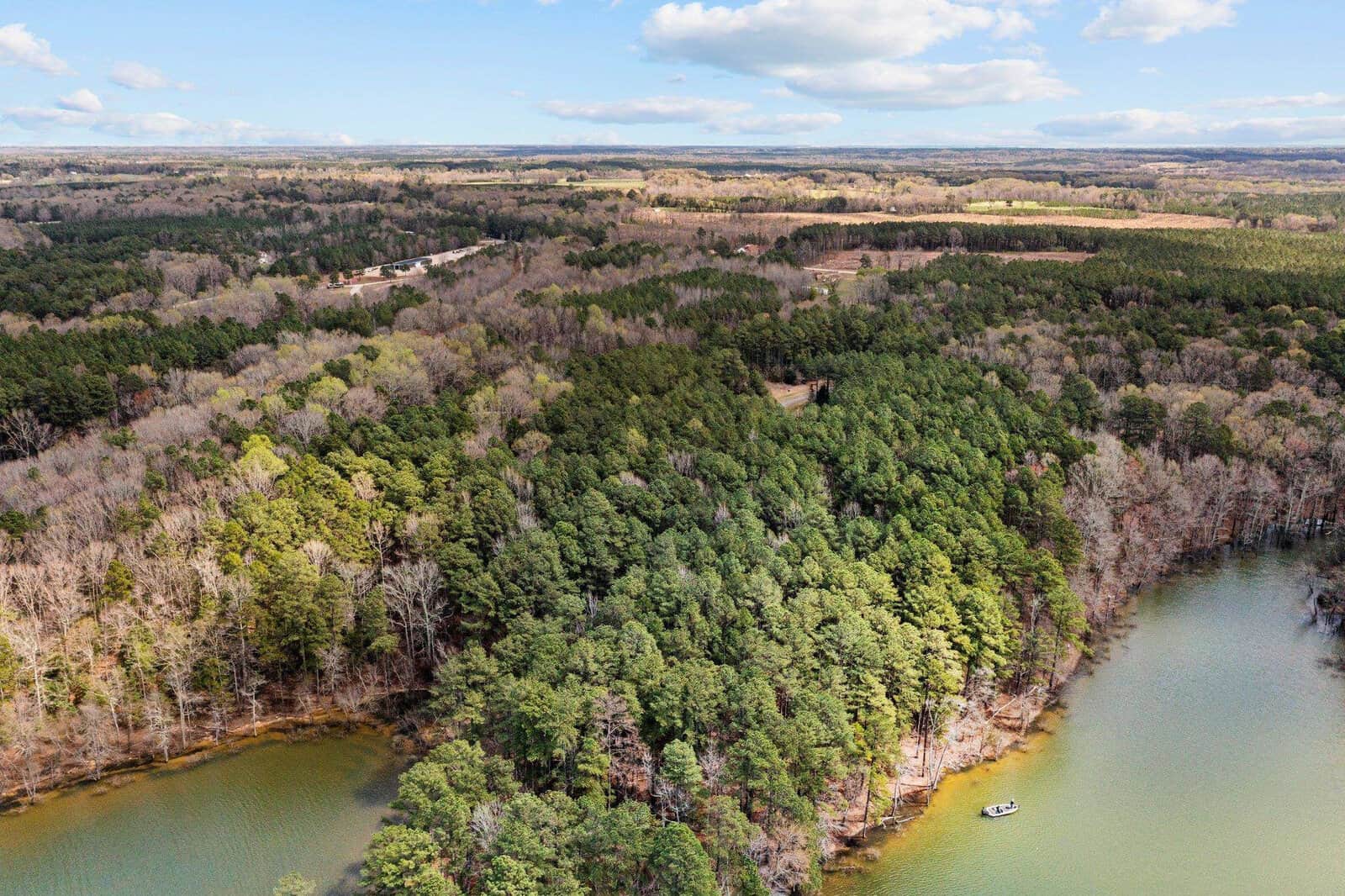 Aerial view of a forested area with a winding shoreline and a small boat on greenish water. The landscape includes mixed trees, open fields, and a blue sky with scattered clouds in the background.