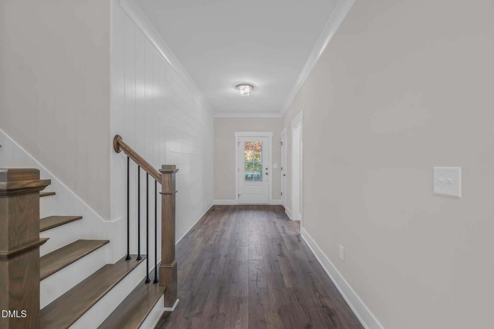 A hallway with wood flooring, white walls, a ceiling light fixture, a staircase with wooden steps and black railings on the left, and a glass door at the end of the hall leading outside.