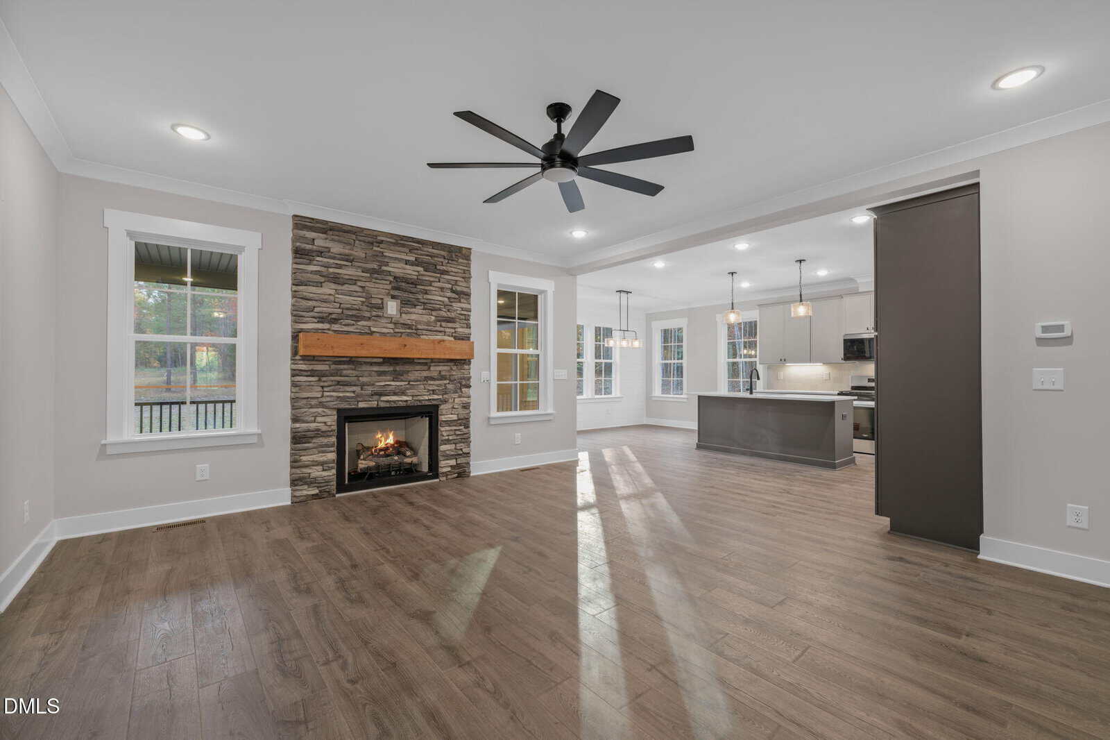 Open living area with wood floors, a ceiling fan, and a stone fireplace. The space connects to a modern kitchen with an island and pendant lights. Large windows and glass doors provide natural light.