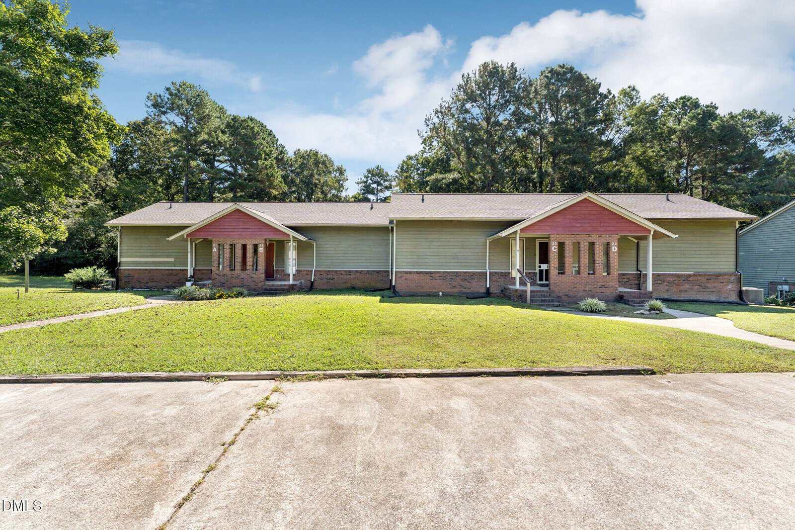 Single-story duplex with green siding and red gable accents, brick foundation, and covered front porches. Well-maintained lawn in front, surrounded by trees, with a concrete driveway in the foreground.