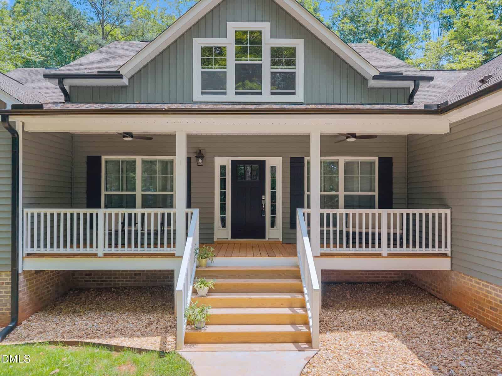 A front view of a modern gray house with white trim, a covered porch, black shutters, double front doors, and a set of wooden steps leading up from a stone pathway. Trees are visible in the background.