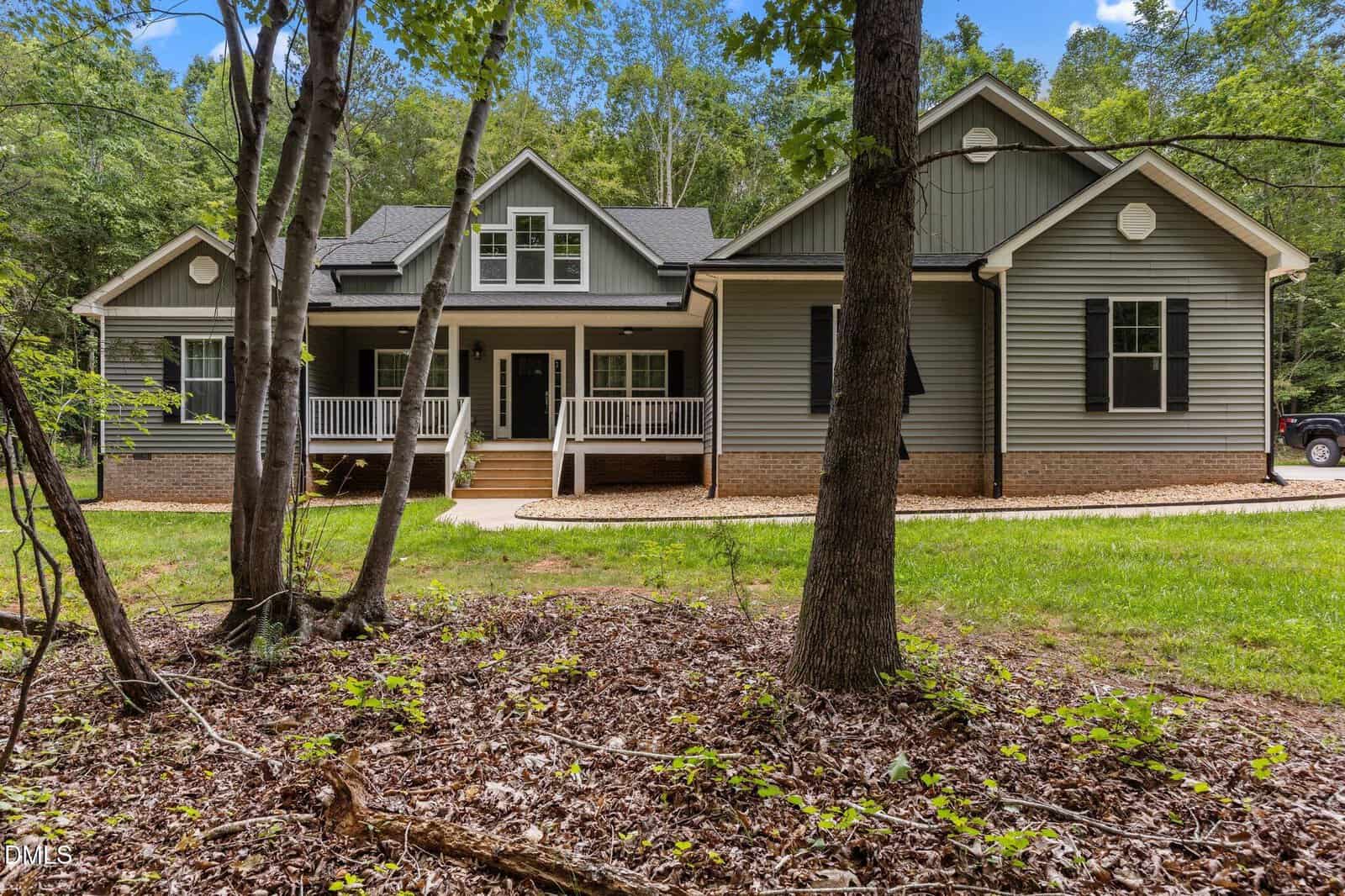 A gray single-story house with white trim, a covered front porch, and brick foundation sits in a wooded area with green grass and trees in the foreground.