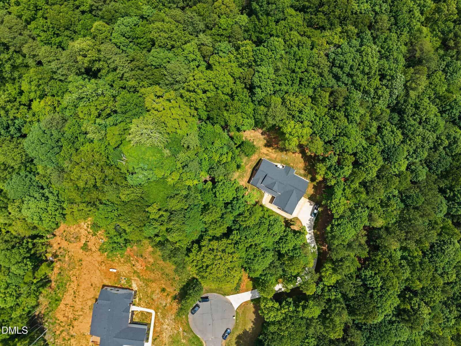 Aerial view of a house surrounded by dense green forest, with another house partially visible nearby and a cul-de-sac at the bottom of the image. Paths and cleared areas can be seen among the trees.