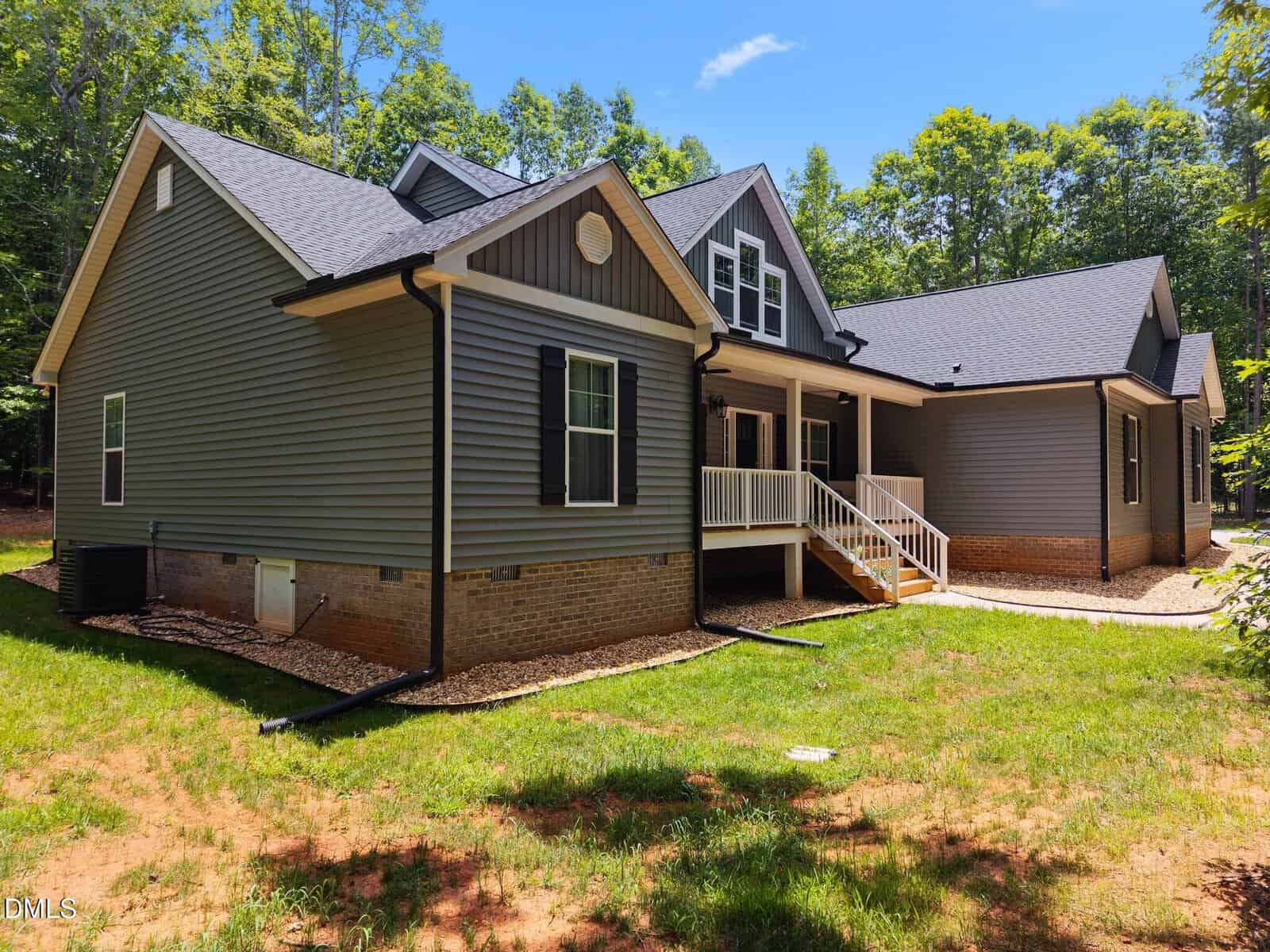 Single-story house with gray siding, black shutters, and a gabled roof. The home has a covered front porch with steps, white railings, brick foundation, and is surrounded by trees and a grassy yard.
