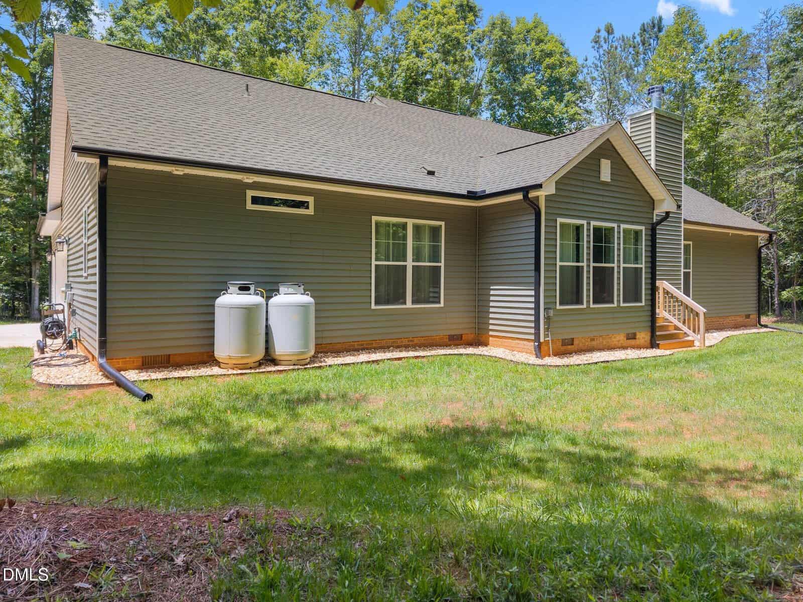 A single-story gray house with a dark roof, two propane tanks on the side, a small porch with steps, and surrounded by trees and grass on a sunny day.