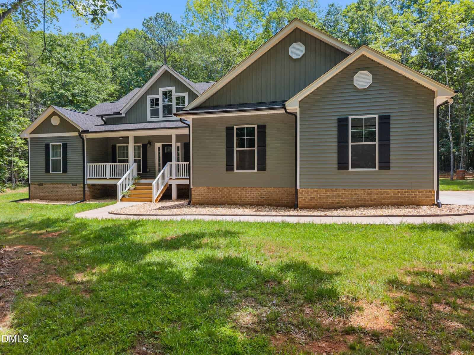 A charming single-story house at 5300 Langdon Drive, Oxford, with gray siding, white trim, black shutters, and a brick foundation. Enjoy the covered front porch surrounded by trees and lush green grass.