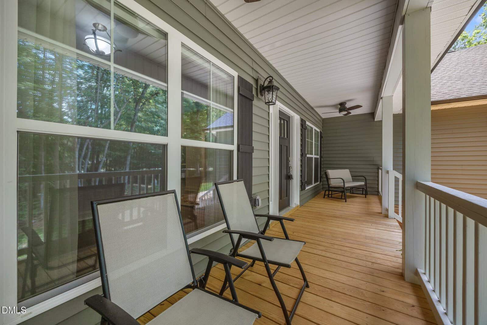 A covered porch with a wooden floor features three metal and mesh chairs, large windows on one side, and white railings. Ceiling fans and lights are mounted overhead. Another house is partially visible next door.