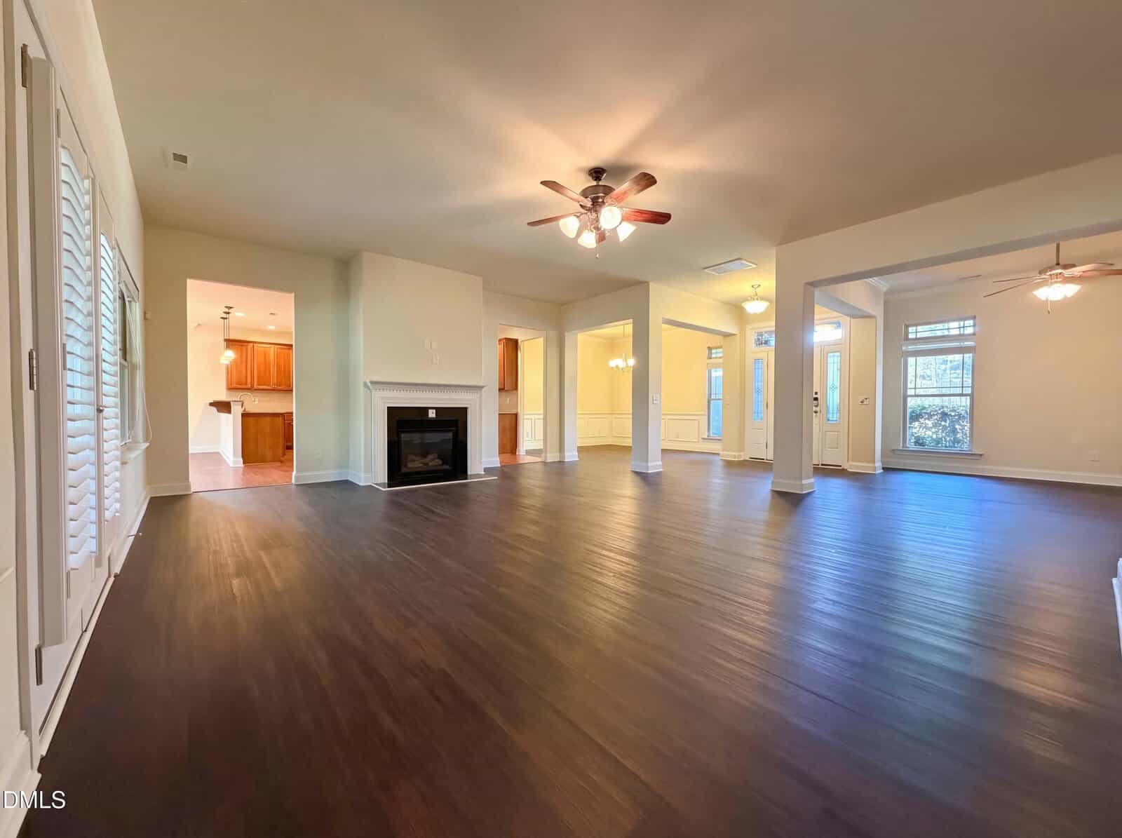 Spacious empty living room with dark wood floors, a ceiling fan with lights, a white fireplace, and an open layout leading to a kitchen and dining area. Large windows and glass doors provide natural light.