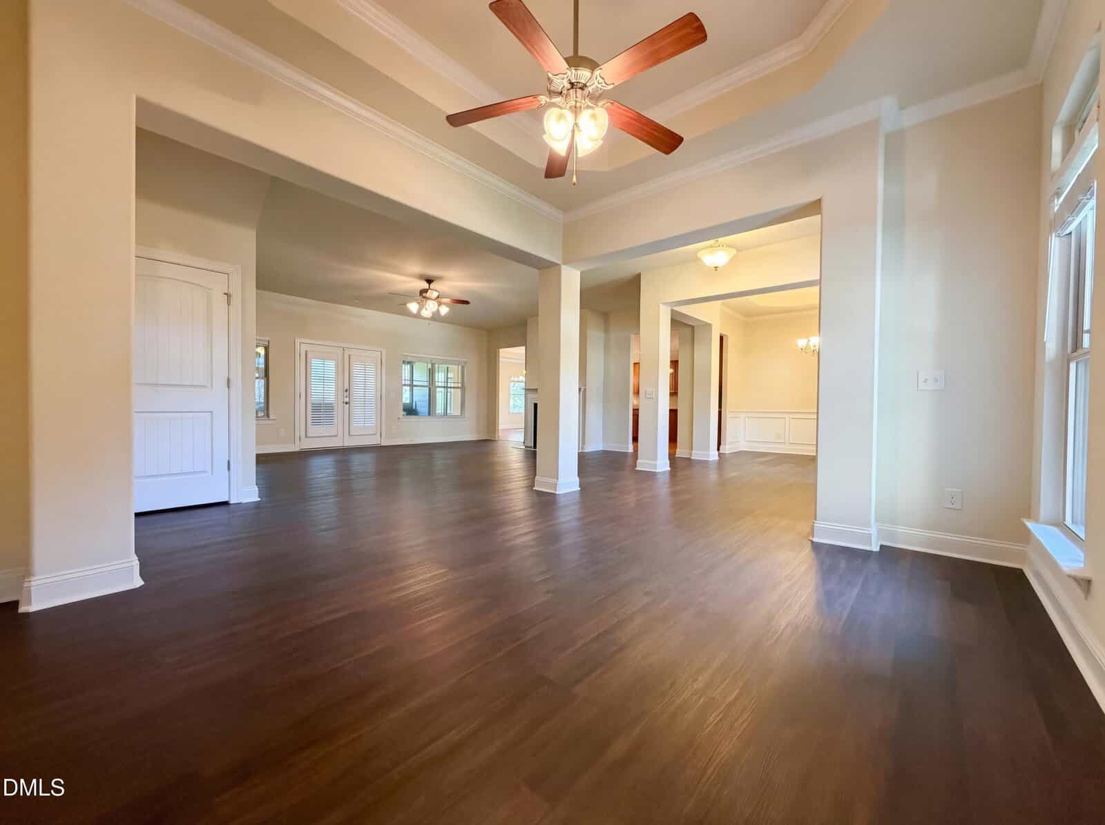 Spacious open-plan living area with dark wood floors, beige walls, large windows, ceiling fans, and multiple light fixtures. White trim and columns add elegance to the modern, empty interior.