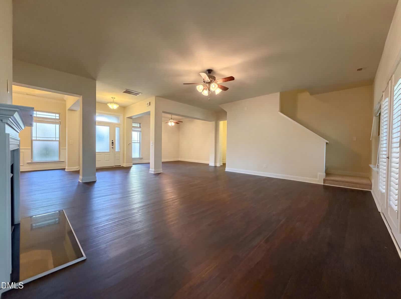 Spacious empty living room with dark hardwood floors, ceiling fans, white walls, fireplace, large windows, and a staircase leading upstairs.