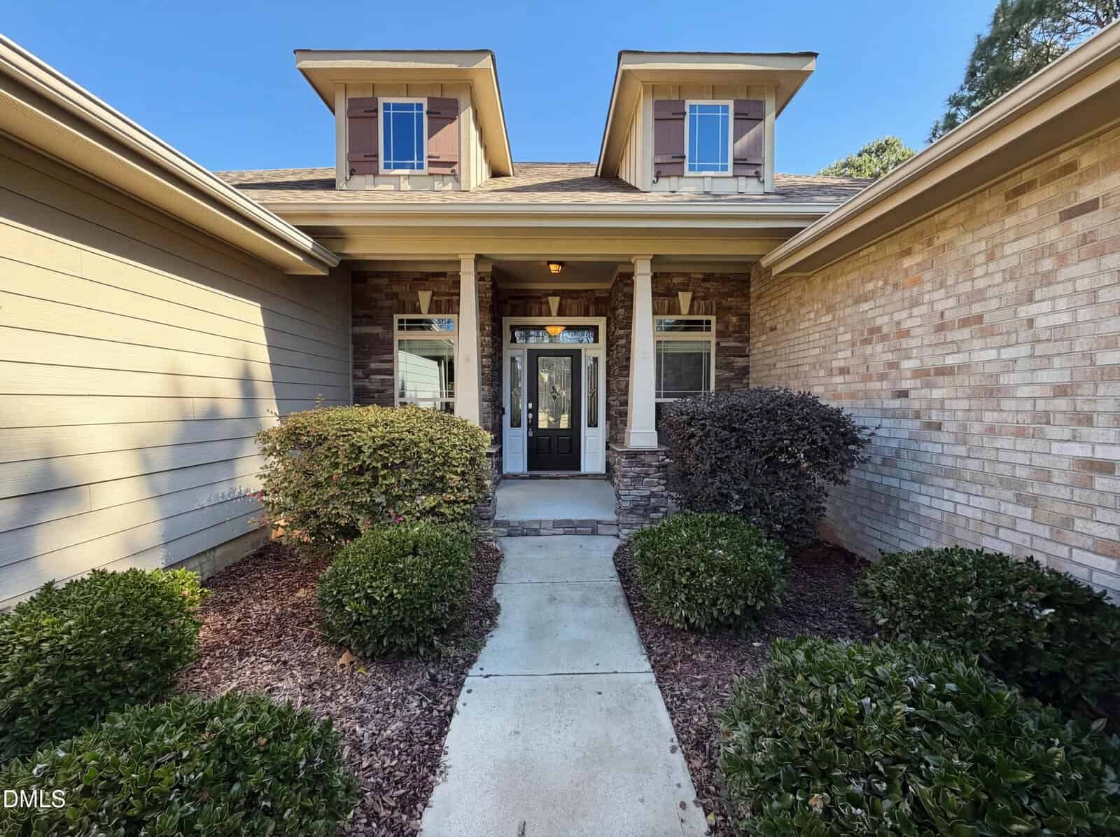 A concrete walkway leads to a covered front porch with two columns, double front doors with glass panels, and dormer windows above; brick and siding exterior with neatly trimmed bushes.