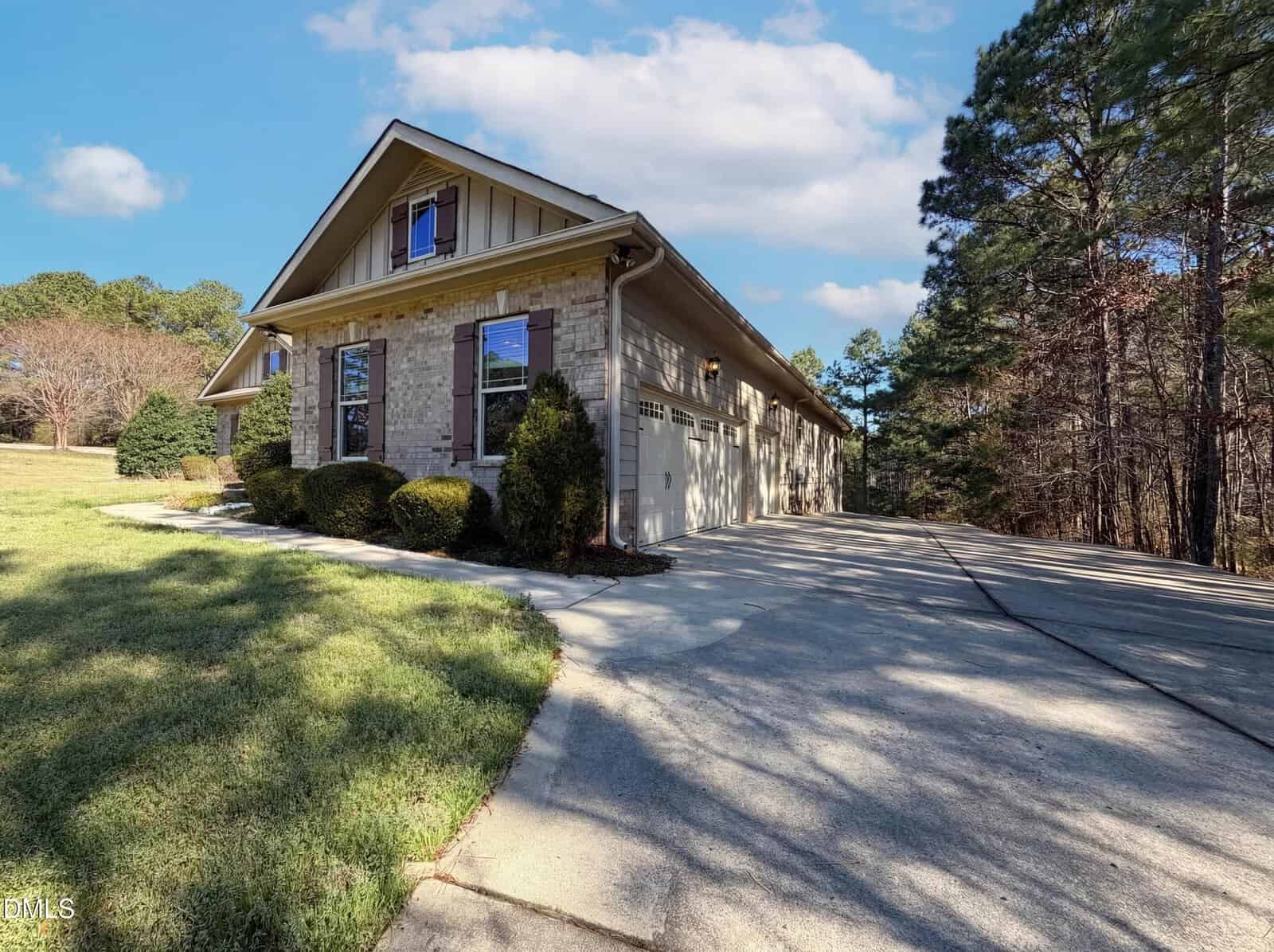 A single-story house with a brick exterior, gable roof, and attached garage. The home is surrounded by grass, shrubs, and trees, with a driveway leading up to the garage under a partly cloudy sky.