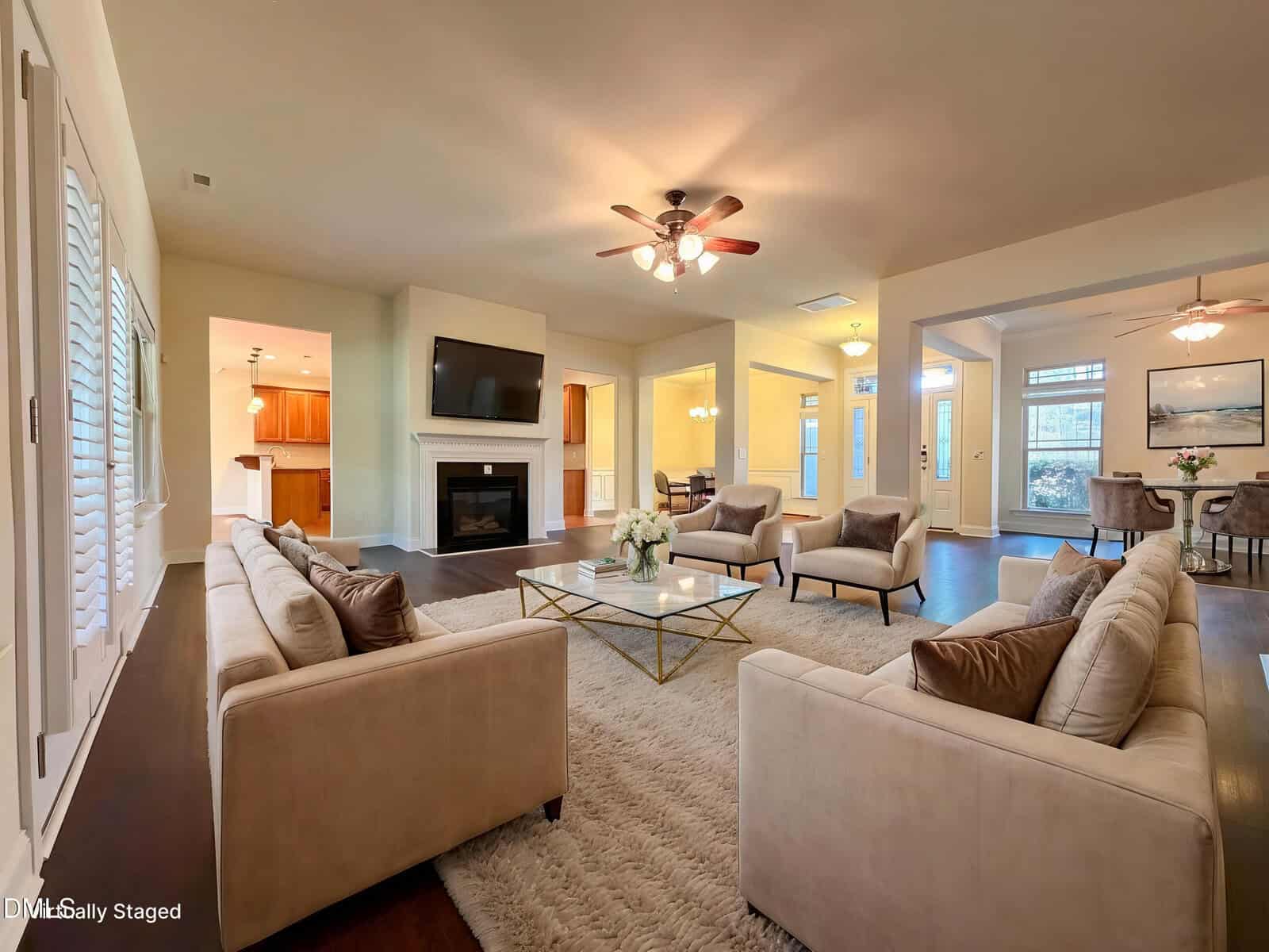 A spacious living room with beige sofas, a white rug, a glass coffee table, and a wall-mounted TV above a fireplace. The open layout leads to a dining area and kitchen, with wooden floors and ceiling fans.