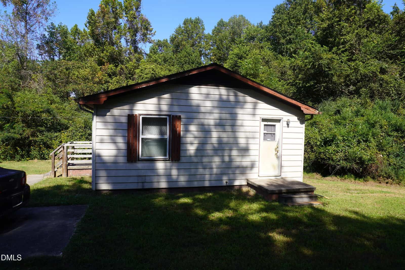 A small, single-story house with light-colored siding, a dark brown roof, and wooden shutters. There is a single window and a door with steps. The house is surrounded by greenery and trees.