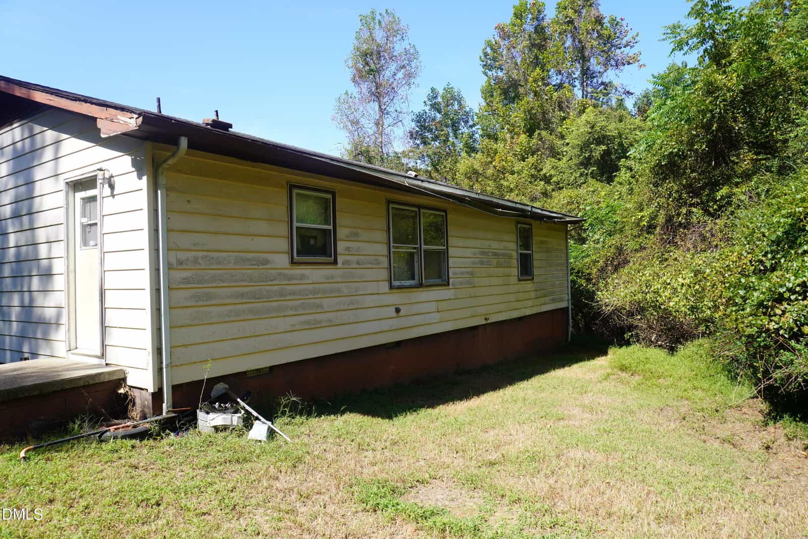 A single-story house with yellow siding and a dark roof shows signs of wear and dirt. Two windows are visible. The yard is grassy and bordered by dense, overgrown bushes and trees under a clear blue sky.