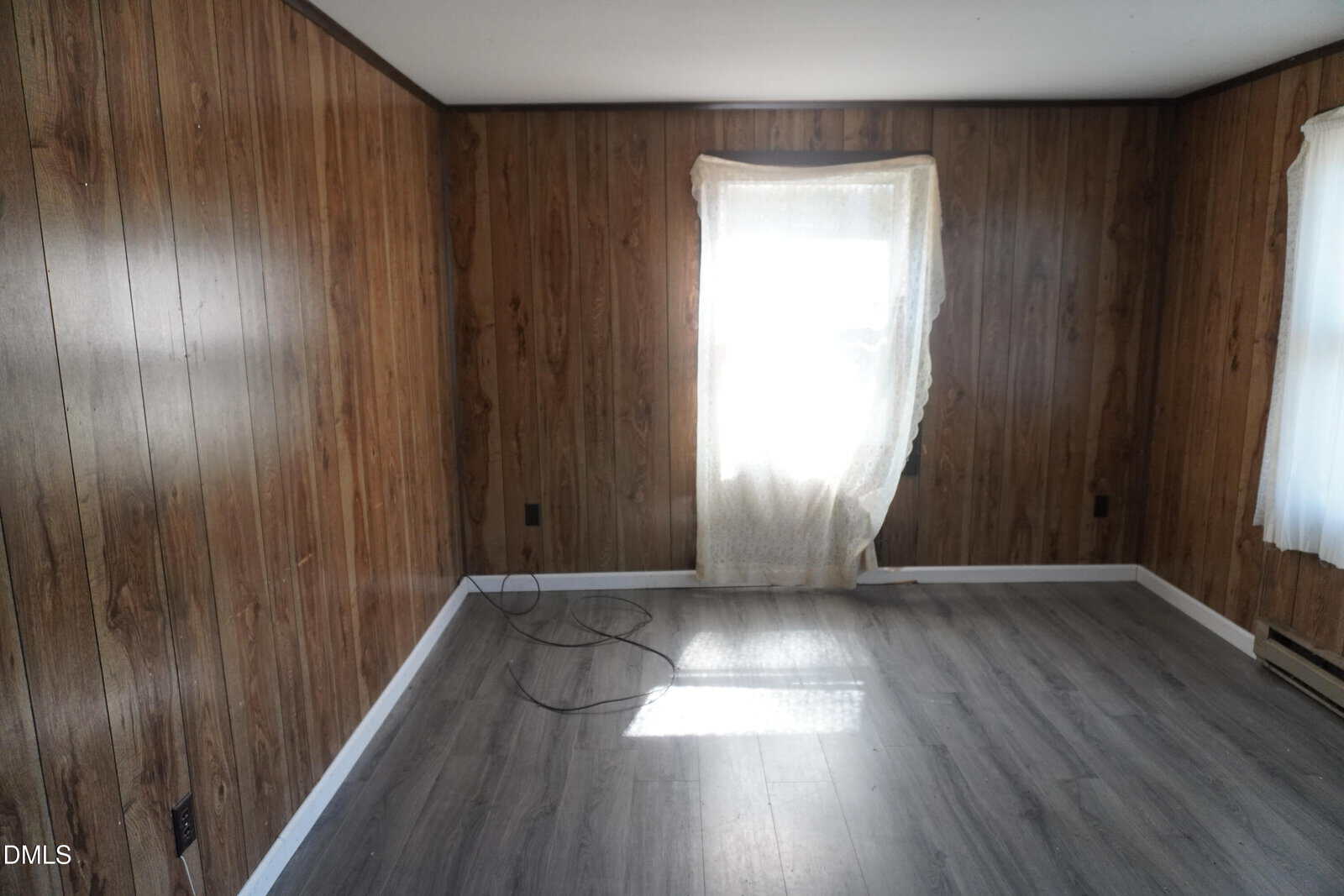 An empty room with wood-paneled walls, gray laminate flooring, a window covered by a sheer curtain letting in sunlight, and loose black cables on the floor near the wall.