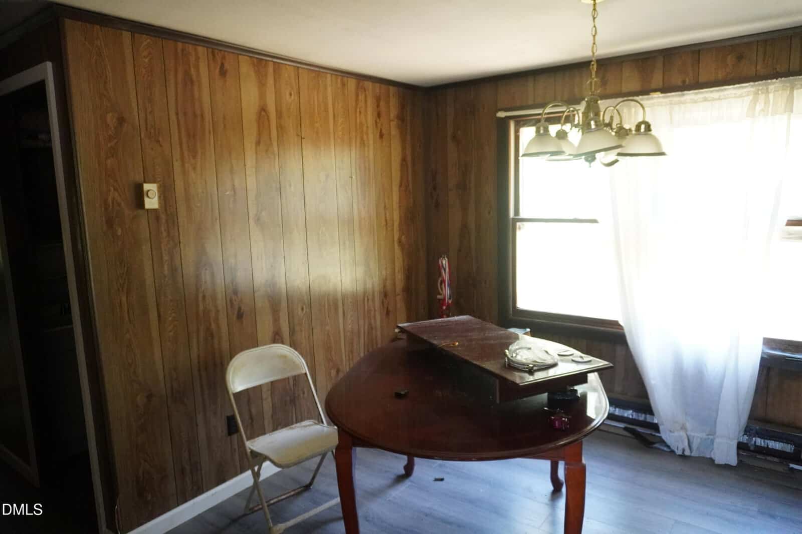 A small room with wood-paneled walls, a round wooden table with items on top, a folded white metal chair, and a window with sheer curtains letting in sunlight. A ceiling light fixture hangs above the table.