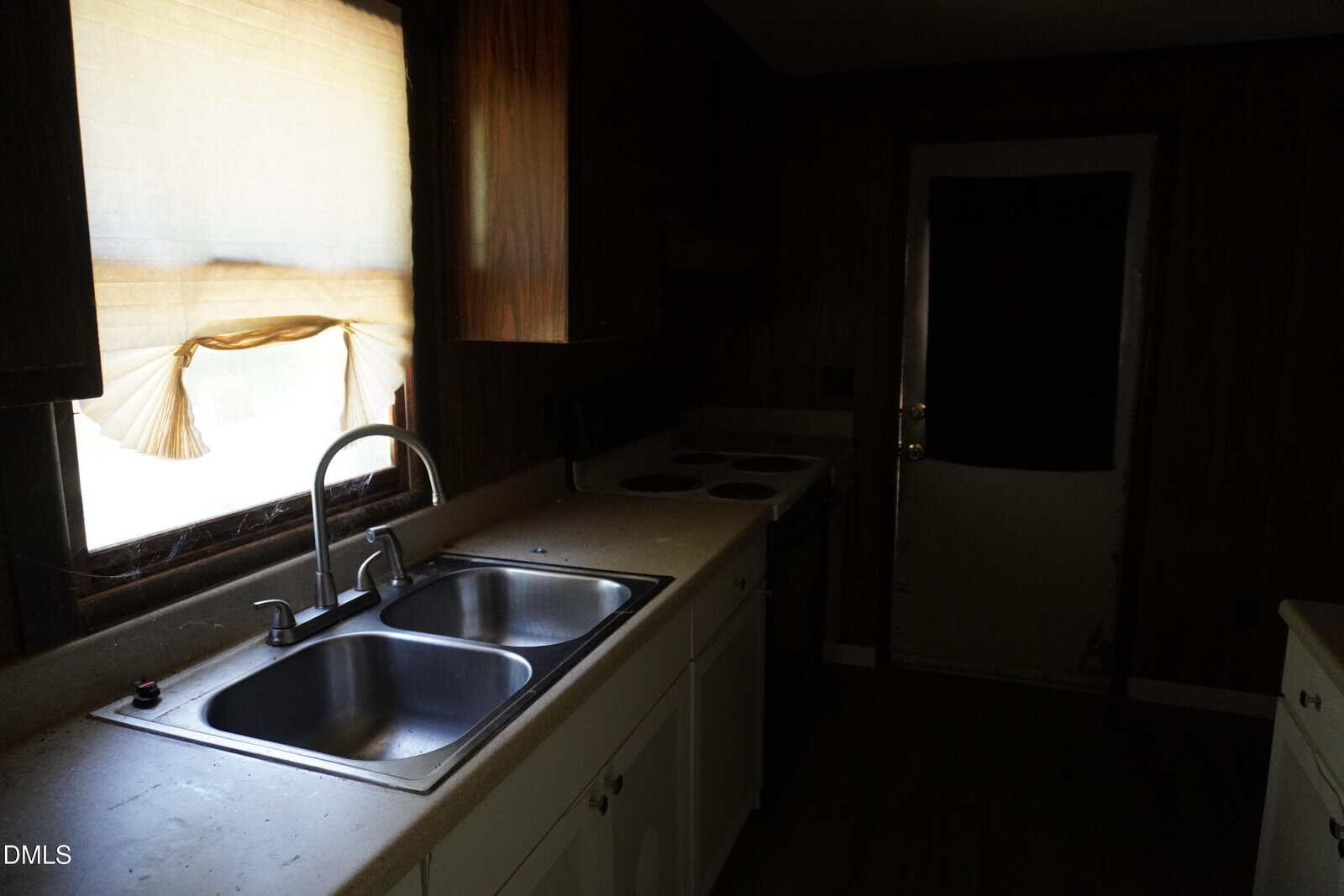 A dimly lit kitchen with a double sink, white cabinets, an electric stove, and a countertop. Sunlight comes through a window with a torn curtain. A closed door is visible in the background.