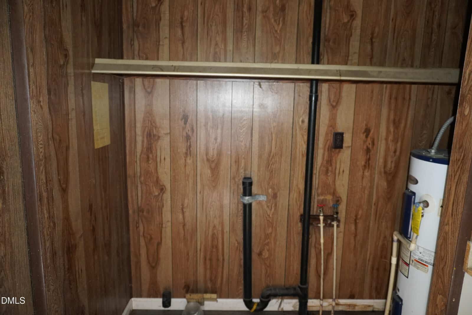 A small utility room with wood-paneled walls, exposed plumbing pipes, a water heater on the right, and a horizontal wooden board mounted above the pipes. A paper is attached to the left wall.