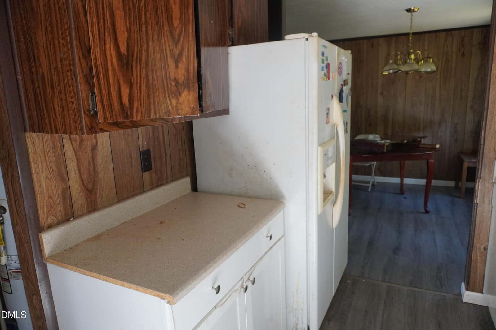 A kitchen with wood-paneled walls, stained countertop, and a white refrigerator with visible dirt. A dining area with a table and gold chandelier is partially visible in the background.