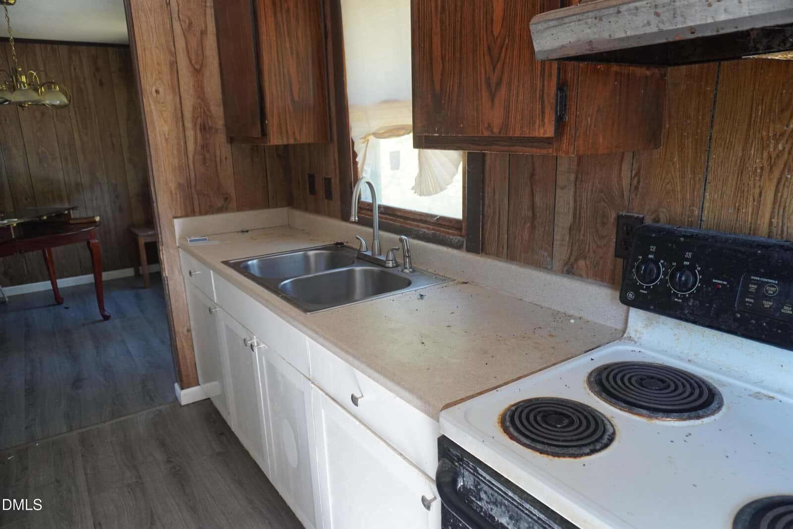 A kitchen with wood-paneled walls, upper wooden cabinets, lower white cabinets, a double sink, and an electric stove with four burners. The countertop shows some dirt and stains. A dining area is visible in the background.