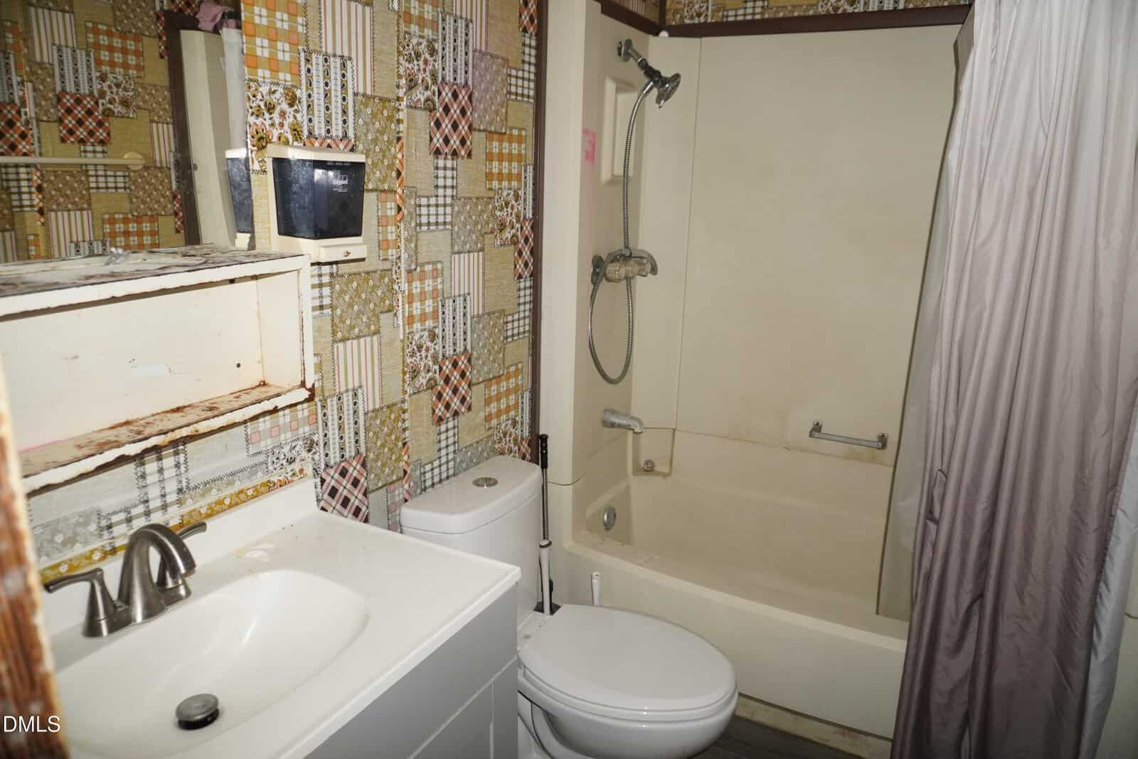 A small bathroom with patterned wallpaper, a white sink with a faucet, a toilet, and a cream-colored bathtub with a showerhead and grab bar. The wall shelf above the sink has visible rust and wear.
