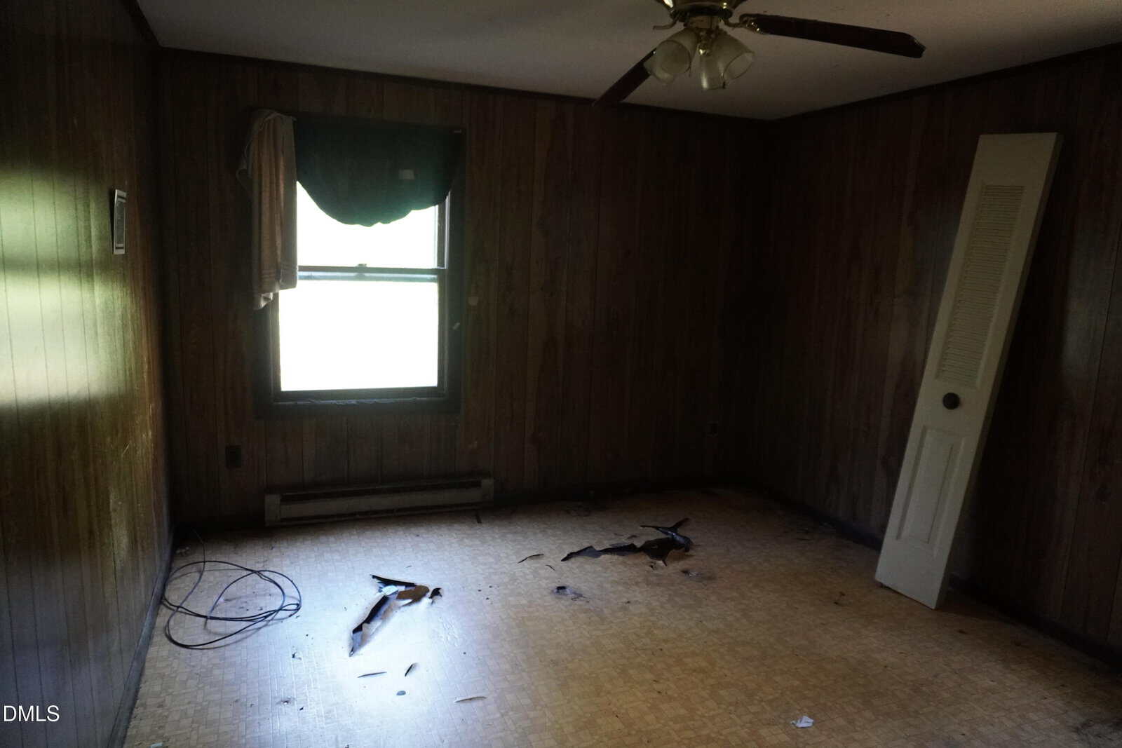 A dimly lit, empty room with wood-paneled walls, a broken window shade, a ceiling fan, a detached door leaning against the wall, debris and wiring on the floor, and peeling linoleum flooring.