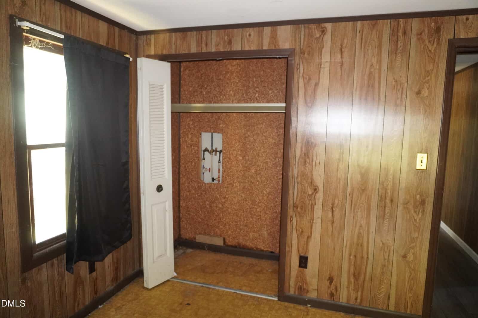 Room with wood-paneled walls, a closet with a missing door, a window with a black curtain, and a small wall mirror inside the closet. The floor has a light brown linoleum pattern.
