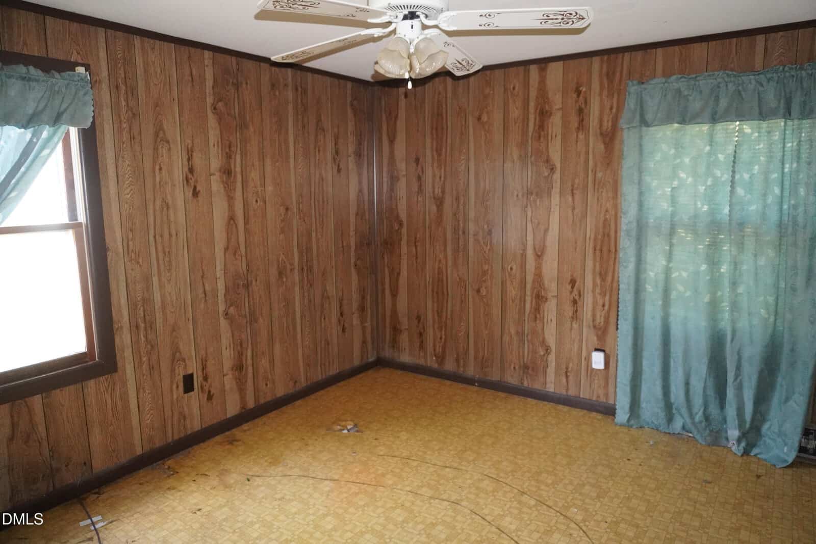 Empty room with wood-paneled walls, yellow patterned flooring, a white ceiling fan, two windows with teal curtains, and some small debris on the floor.
