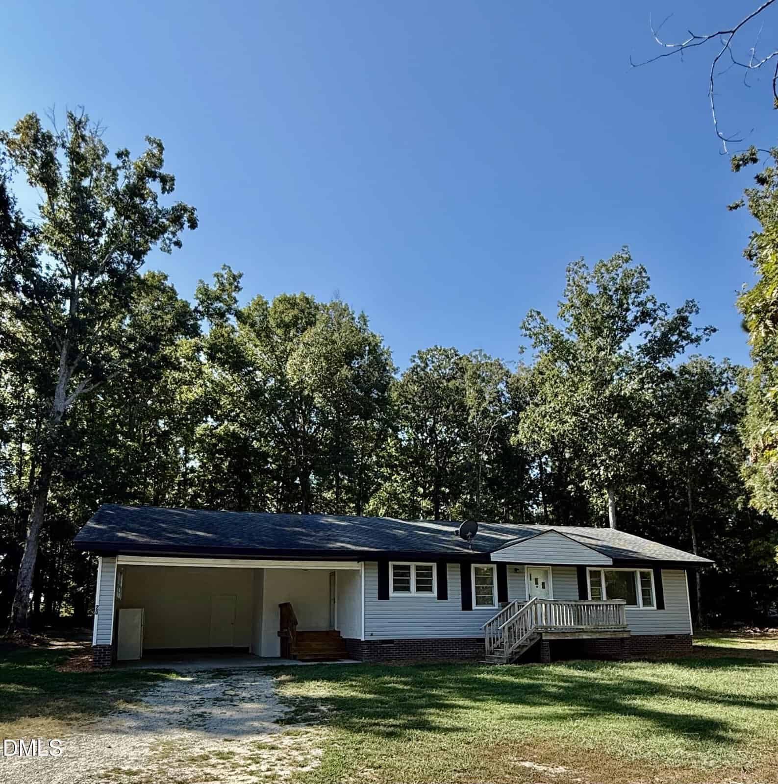 Single-story house with a front porch, carport, and white siding with brick trim, set among tall trees at 20 Acer Way in Zebulon. The sky is clear and blue, and the yard has patchy grass and a dirt driveway.
