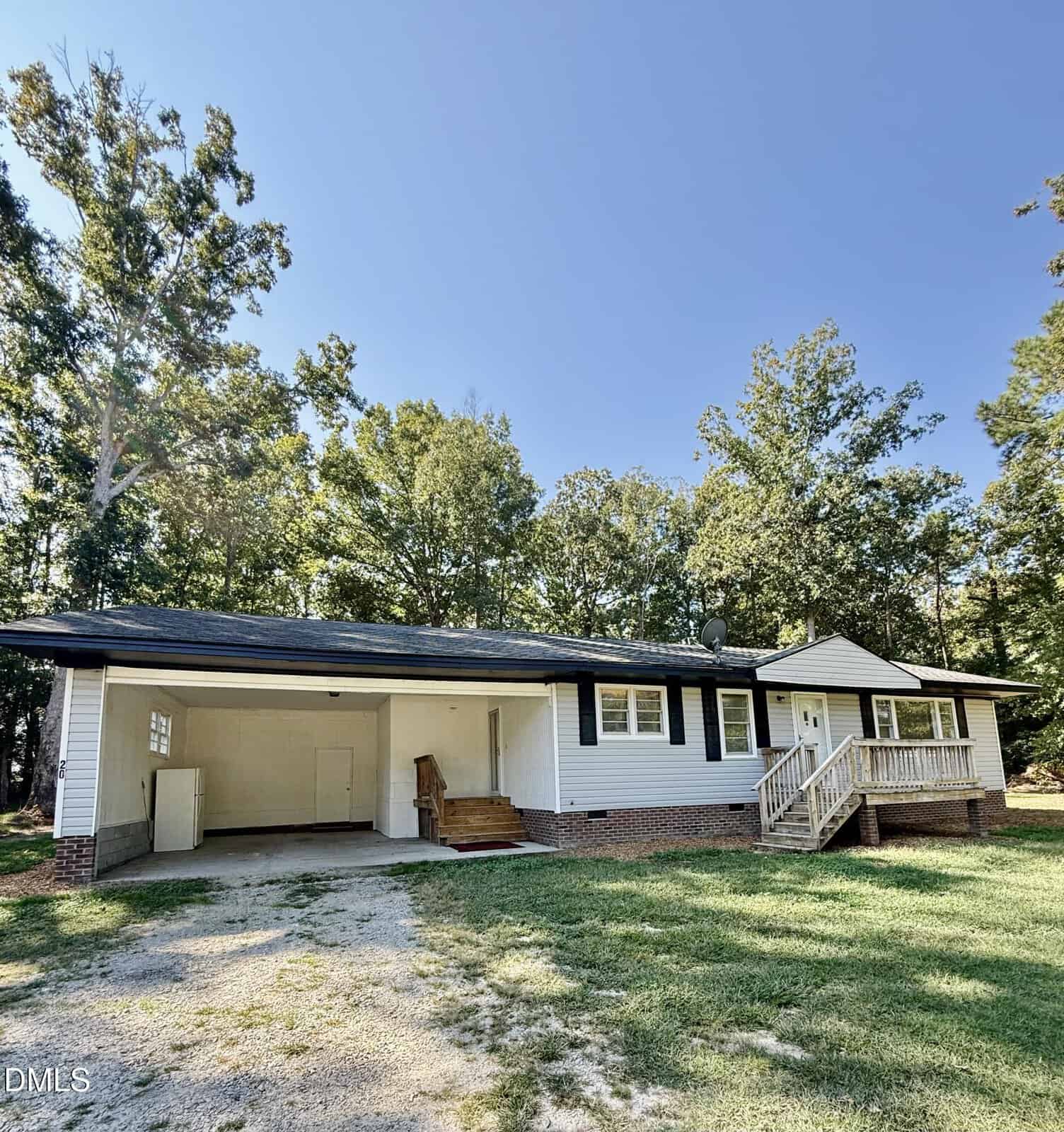 Single-story house with white siding, black shutters, and a brick foundation. Features an attached carport on the left, a front porch with steps, and a lawn bordered by trees under a clear sky.