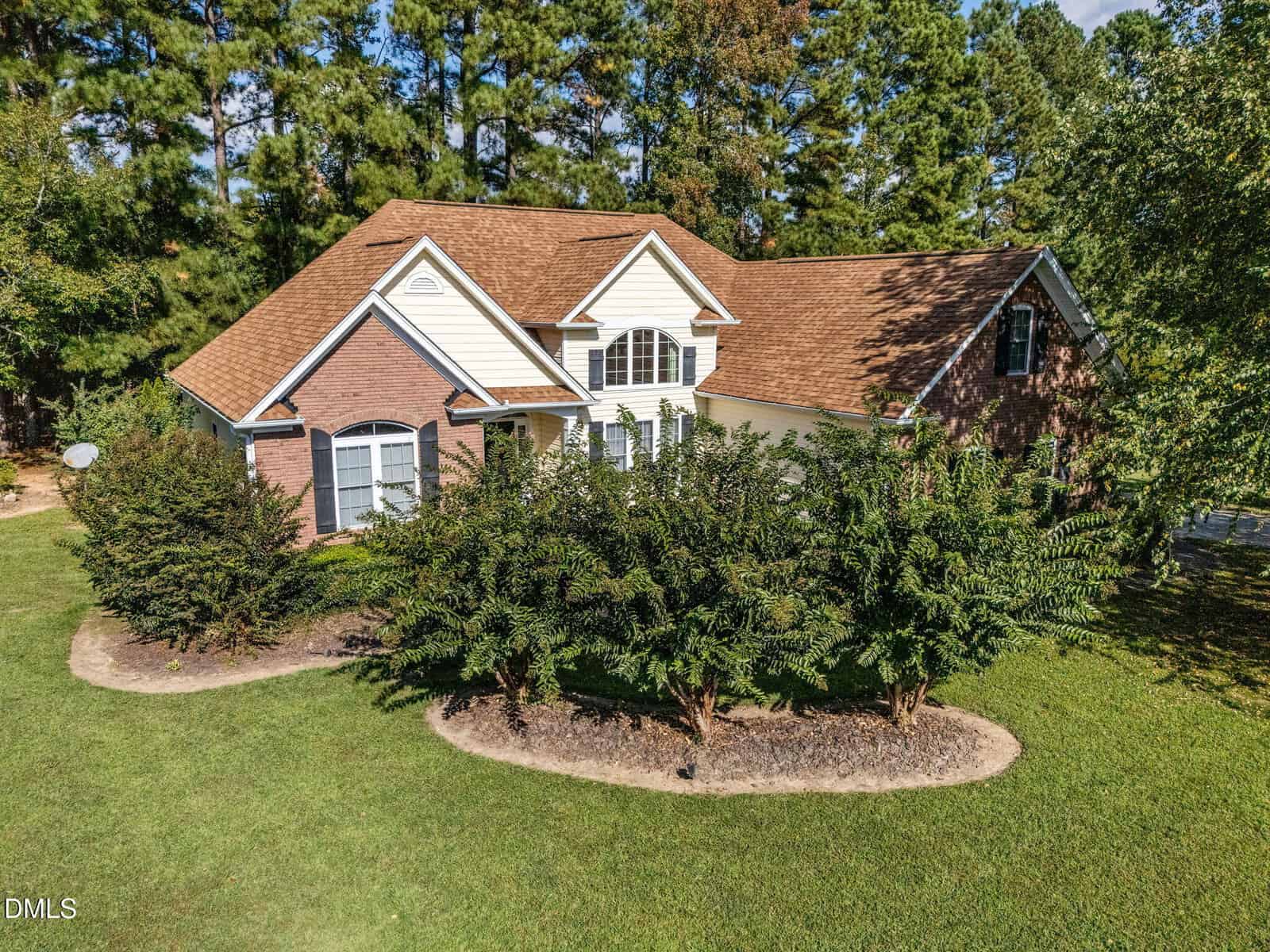 A two-story house with a brown shingle roof, white siding, and brick exterior sits behind a row of trimmed bushes on a green lawn, surrounded by tall trees under a partly cloudy sky.