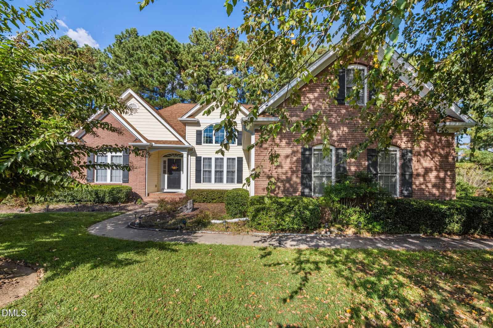 A two-story suburban house with red brick and white siding sits at 3538 Saddle Ridge Road, surrounded by green grass, manicured bushes, and trees under a blue sky with scattered clouds.